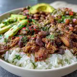 Golden-brown shredded carnitas sit atop fluffy white rice with pinto beans, fresh salsa, and avocado slices.