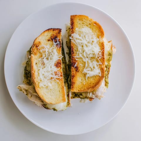 Golden-brown sourdough grilled cheese stuffed with shredded chicken, vibrant pesto, and melted mozzarella, cut in half on a wooden cutting board.  