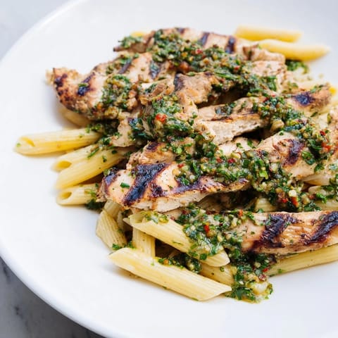 Grilled chicken breast slices and penne pasta coated in vibrant, herby chimichurri sauce on a rustic plate.  
